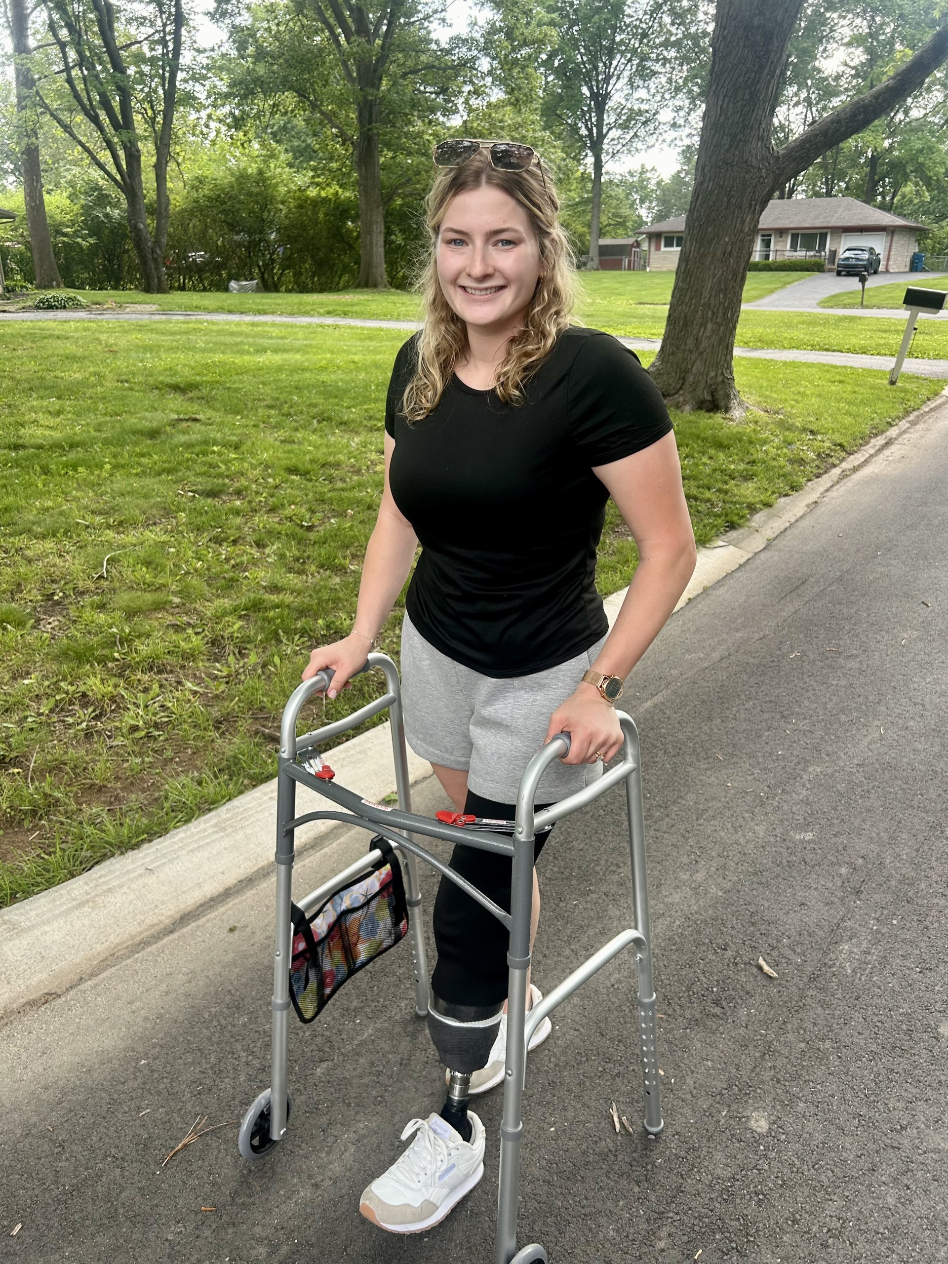 A smiling young woman with curly hair wearing a black shirt and gray shorts, using a walker and standing on a paved path in a grassy, tree-lined neighborhood.