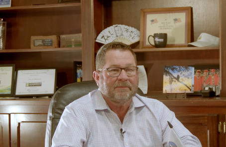 A middle-aged man with a beard and glasses sitting at a desk in an office setting, surrounded by framed photos and other decor.