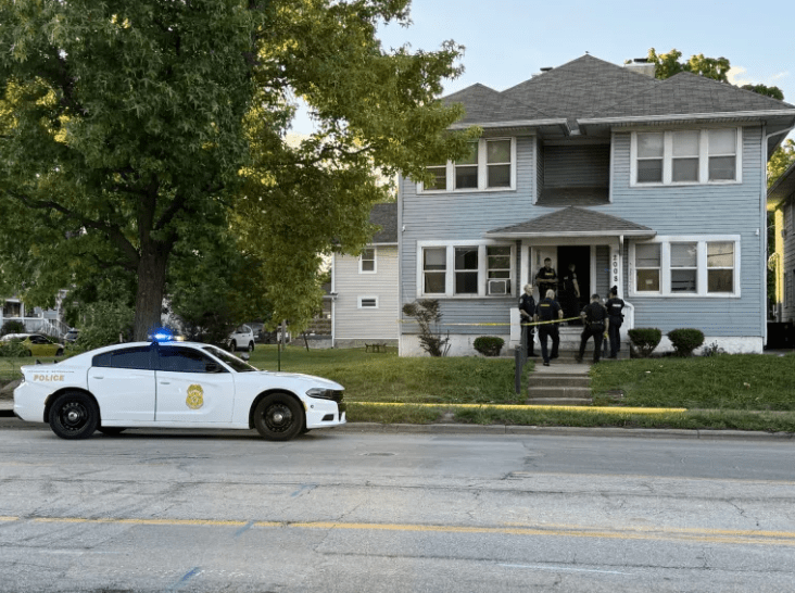 A police car parked on the street outside a residential house, with several police officers standing on the porch.
