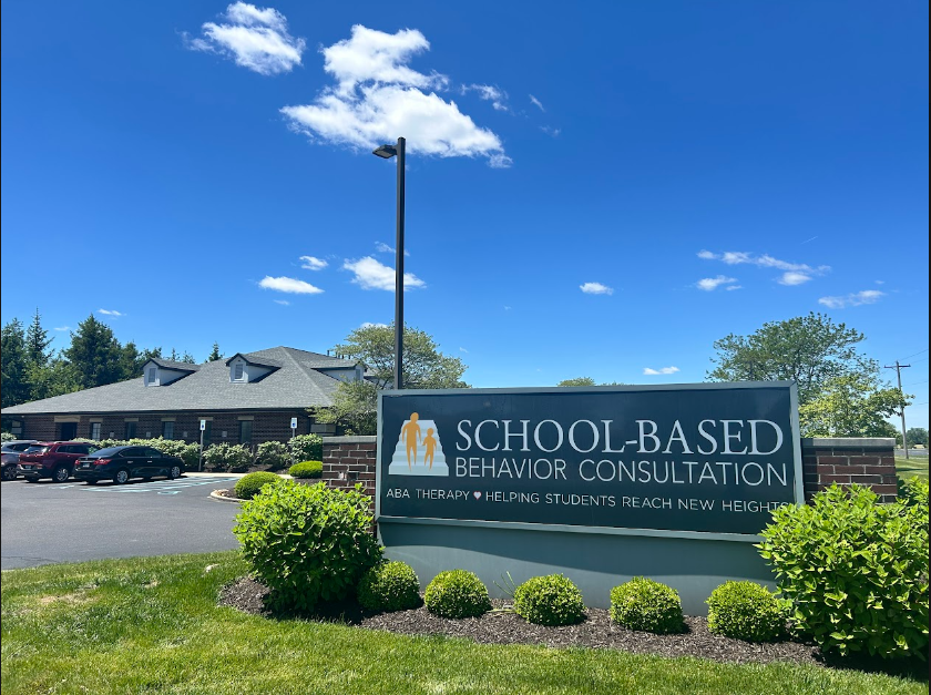 A sign for a school-based behavior consultation service, with the text "SCHOOL-BASED BEHAVIOR CONSULTATION ABA THERAPY • HELPING STUDENTS REACH NEW HEIGHTS" displayed on a brick building.