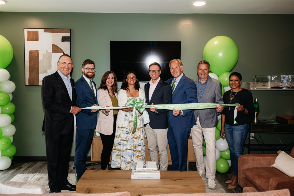 A group of 7 people, including 4 men and 3 women, standing together and holding a large ribbon in front of a green wall with white and green balloons.