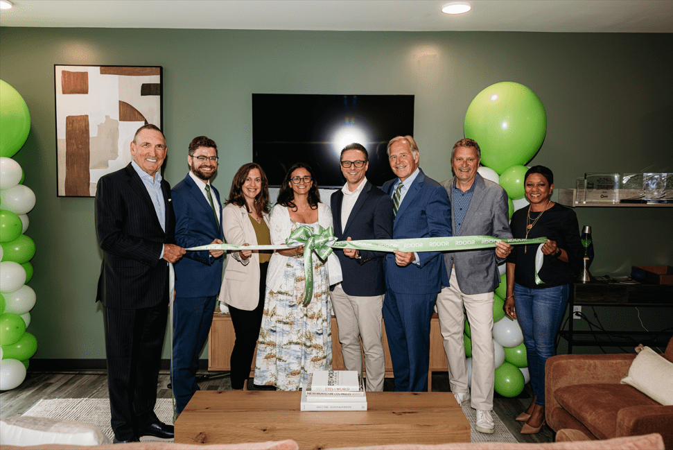 A group of 7 people, including 4 men and 3 women, standing together and holding a large ribbon in front of a green wall with white and green balloons.
