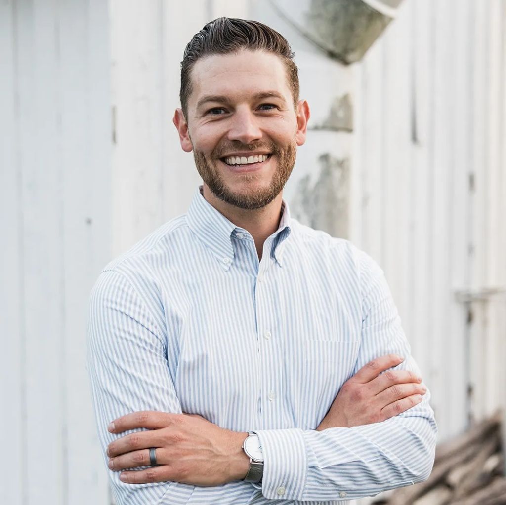 A smiling man with a beard wearing a striped button-down shirt and standing in front of a white wall.