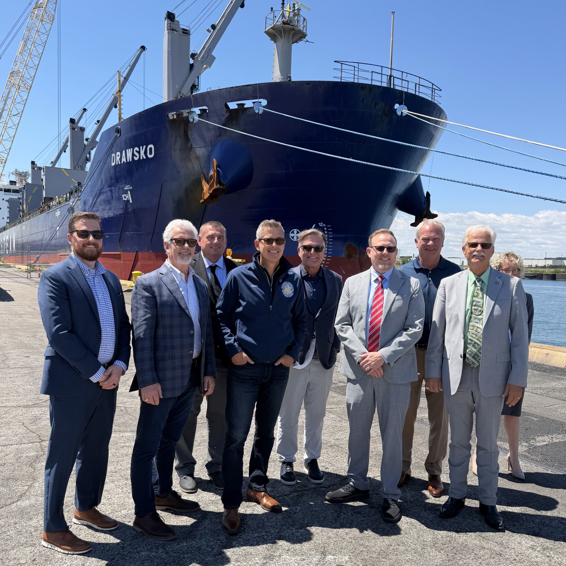 A group of men in suits standing in front of a large blue cargo ship named "DRAWSKO" in a port setting.