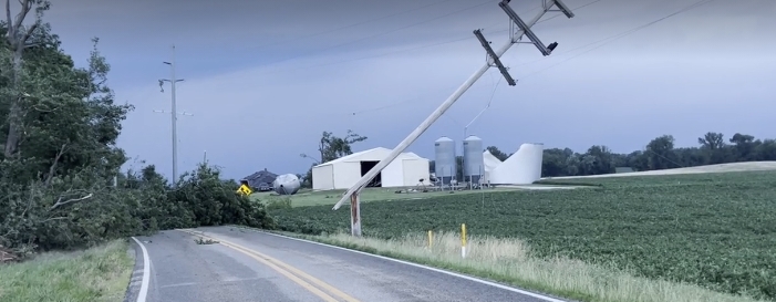 A rural road passing through a grassy field, with a large wind turbine and other industrial structures in the background.