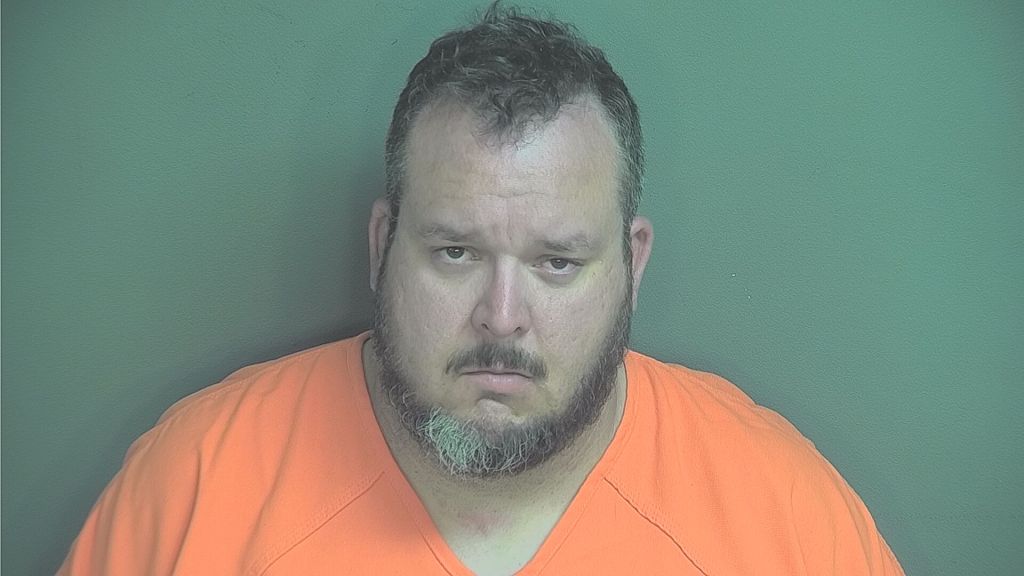 A man with a beard wearing an orange prison uniform looks directly at the camera with a serious expression.
