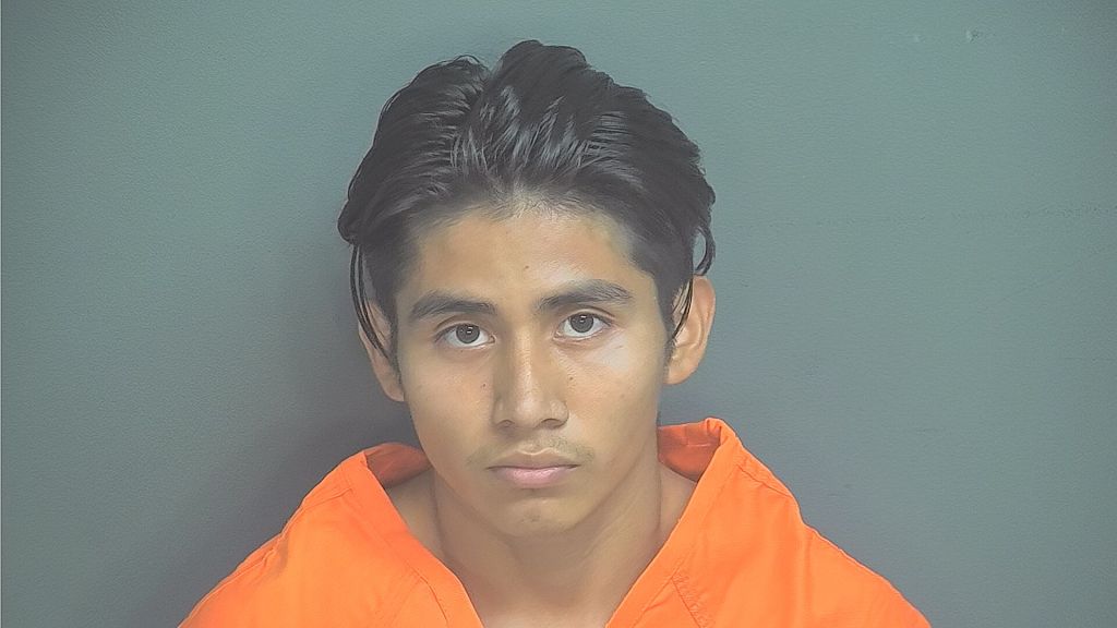 A young man with short dark hair wearing an orange prison uniform looks directly at the camera.