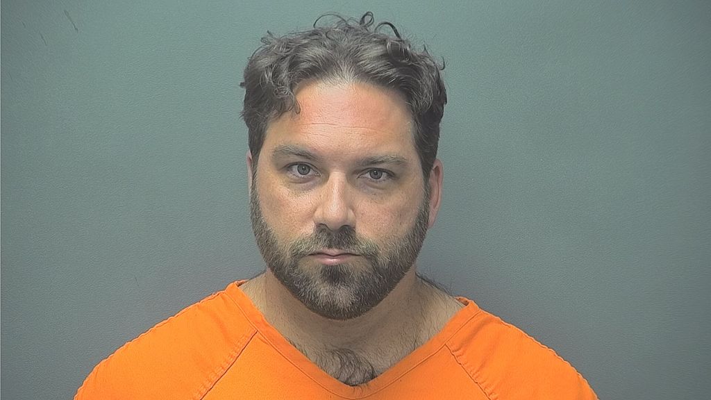 Mugshot of a bearded man with curly hair wearing an orange prison uniform against a gray background.