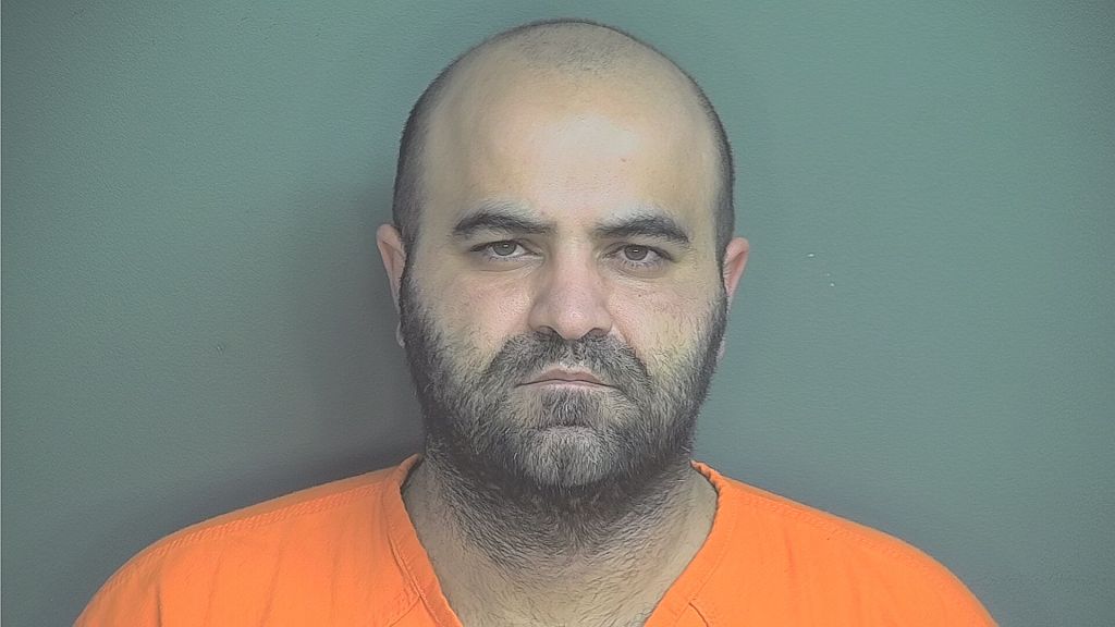 Headshot of a bald, bearded man wearing an orange prison uniform against a green background.