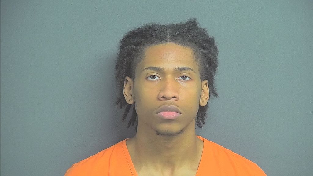 Headshot of a young Black man with curly hair wearing an orange prison uniform against a gray background.