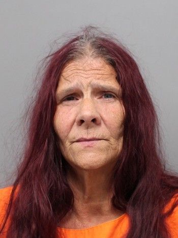 Older woman with long red hair wearing an orange jumpsuit, looking directly at the camera with a serious expression.