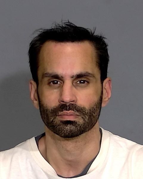 A close-up portrait of a man with dark hair and a beard, wearing a white shirt against a gray background.