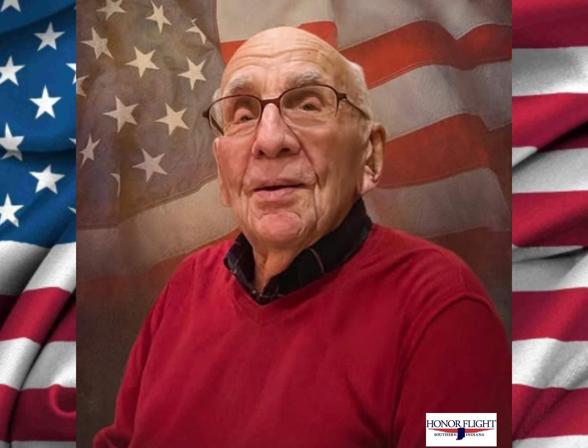 An elderly man with glasses wearing a red sweater, standing in front of an American flag backdrop. The text "HONOR FLIGHT SOUTHERN INDIANA" is visible.