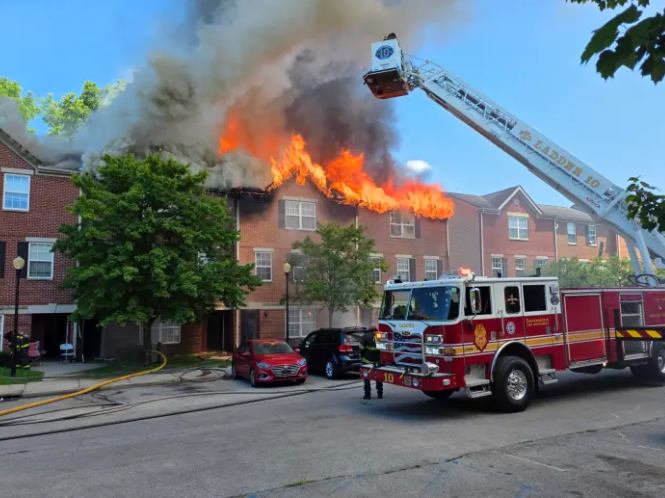 A large fire engulfs a multi-story brick building, with smoke billowing and flames visible. A fire truck with its ladder extended is on the scene.