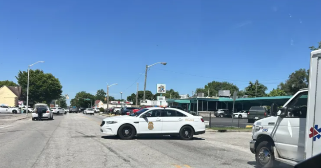 A city street with several vehicles, including a police car, parked along the sidewalk under a blue sky.