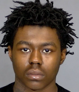 A close-up portrait of a young Black man with dreadlocked hair and a serious expression.