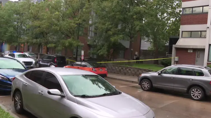 Police tape cordons off an area with parked cars on a street lined with trees.