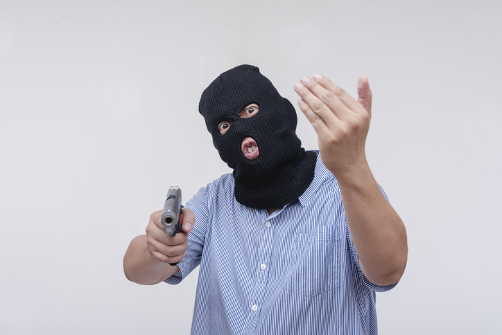 A deranged middle aged man in a black mask threatening someone with a gun to come closer. A robber barking orders to give him money. Isolated on a white backdrop.