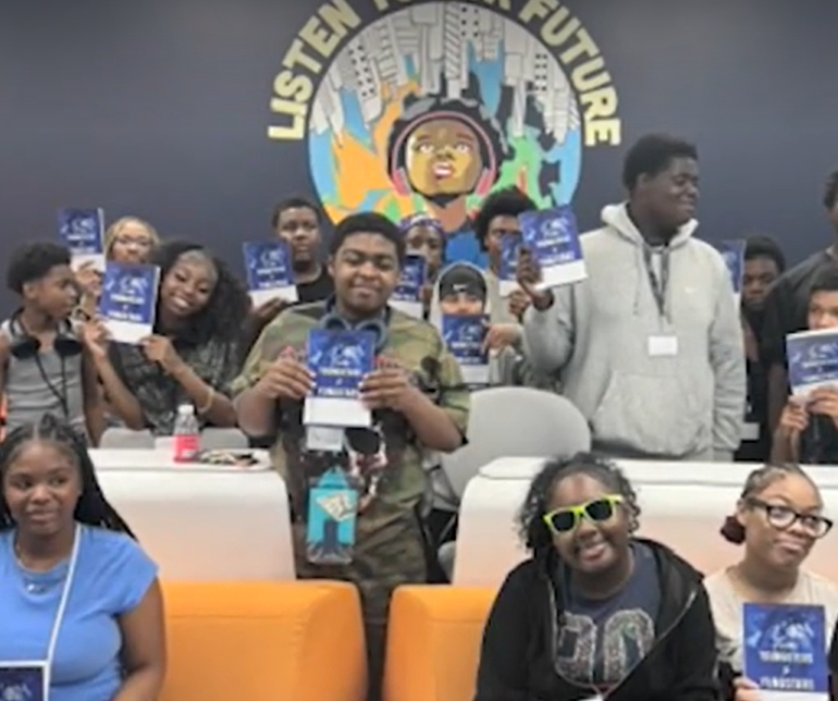 A group of people holding up signs with the "Listen to the Future" logo, gathered in what appears to be a community or event space.