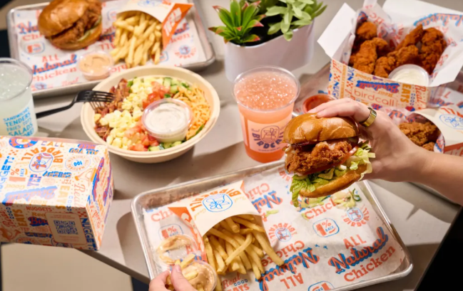 Assortment of fast food items including a burger, fries, and fried chicken, with various sauces and drinks on a tray with colorful branding.