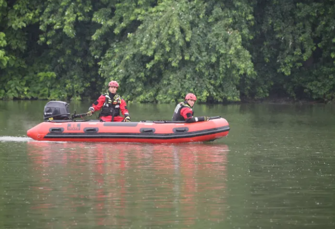 Two people in red life jackets riding in a red inflatable boat on a river surrounded by lush green trees.