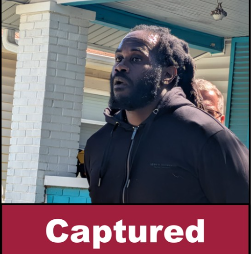 A man with dreadlocks wearing a black jacket stands in front of a brick building with a "Captured" sign.