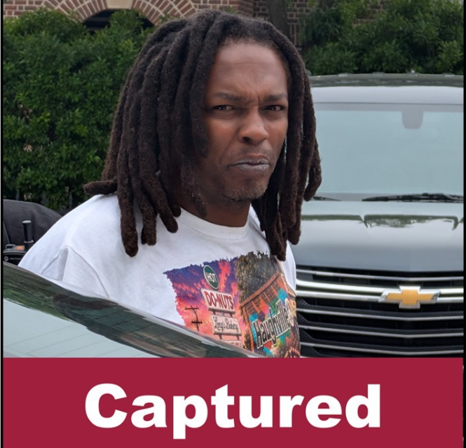 A man with dreadlocks wearing a white t-shirt with a graphic design, standing in front of a Chevrolet vehicle.