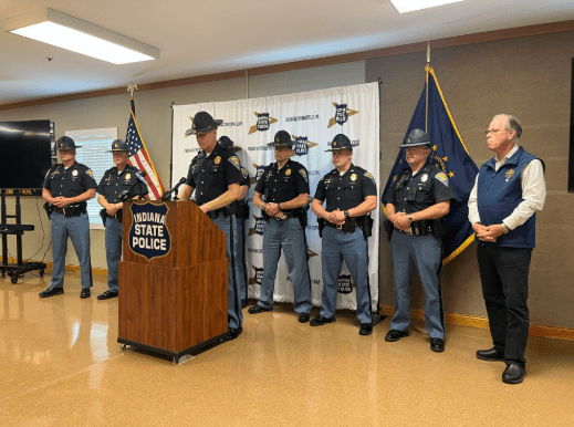 Indiana State Police officers standing in a room, with an Indiana State Police flag and podium visible.