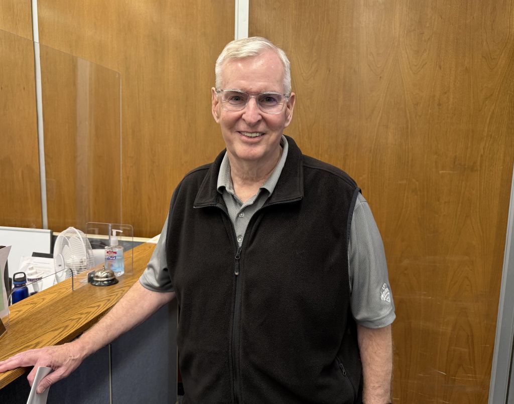 A smiling older man wearing glasses and a black vest stands in an office setting with a wooden desk and sign reading "Tyler".