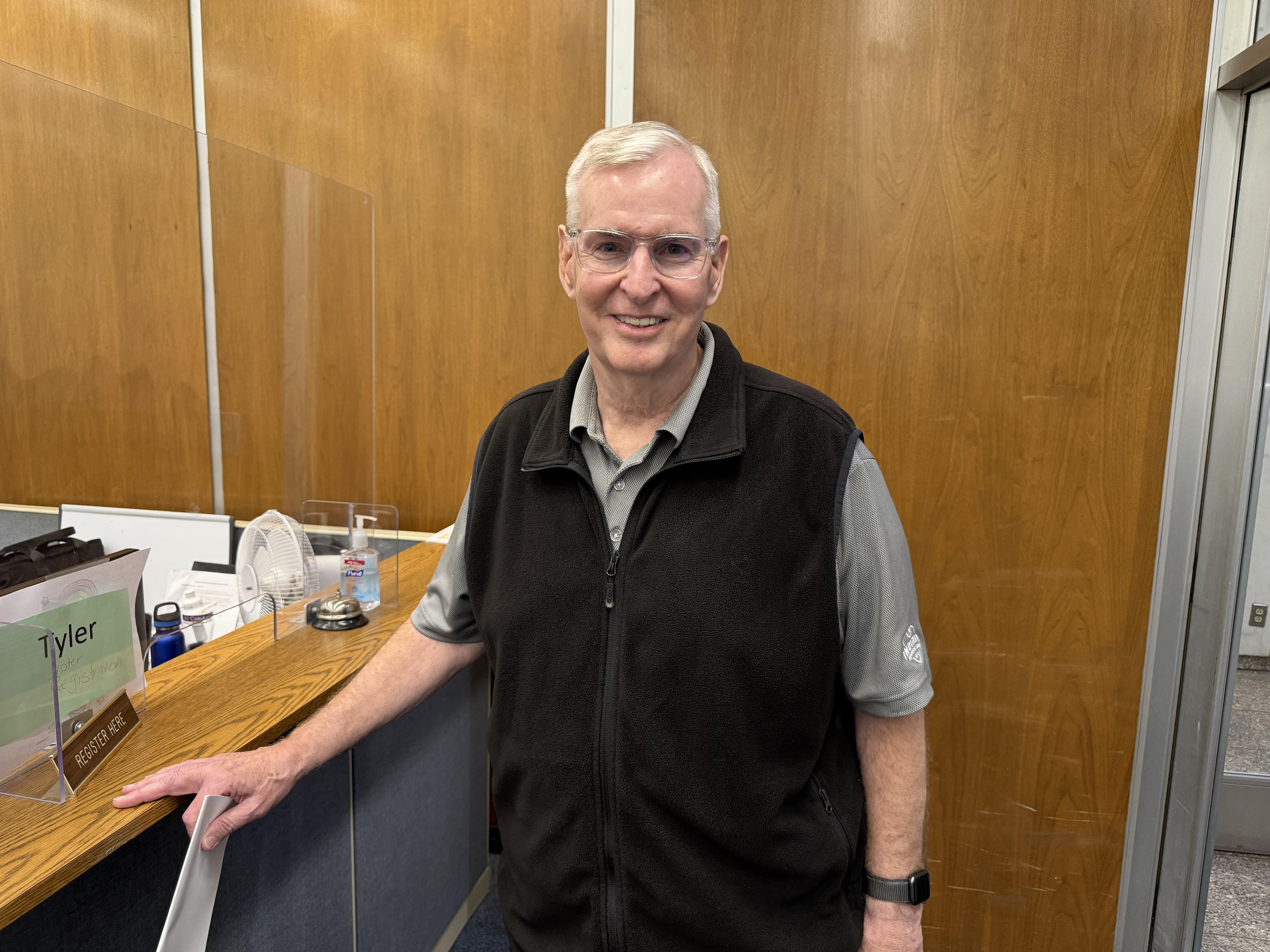 A smiling older man wearing glasses and a black vest stands in an office setting with a wooden desk and sign reading "Tyler".