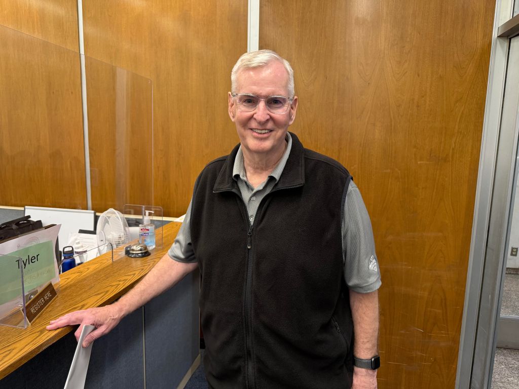 A smiling older man wearing glasses and a black vest stands in an office setting with a wooden desk and sign reading "Tyler".