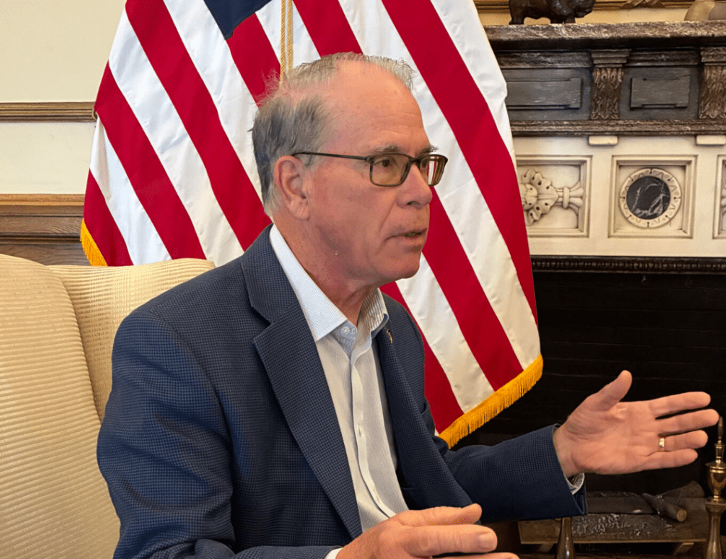 An older man in a suit and glasses speaking at a podium in front of an American flag.