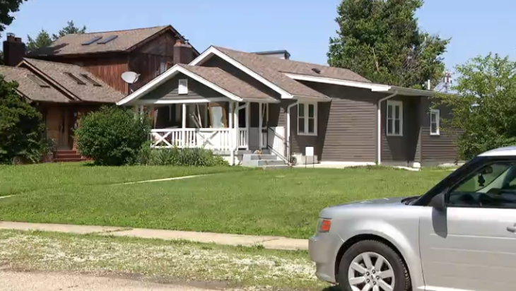 A large two-story wooden house with a porch and a silver car parked in the driveway, surrounded by trees and a grassy lawn.