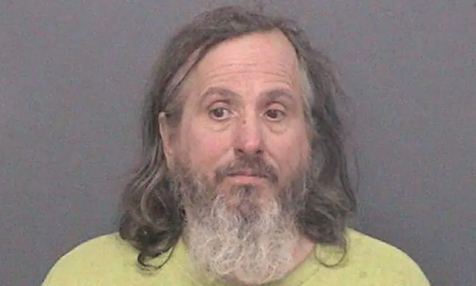 Mugshot of a man with long gray hair and a beard wearing a yellow shirt.