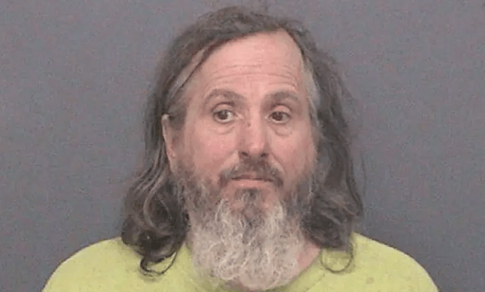 Mugshot of a man with long gray hair and a beard wearing a yellow shirt.
