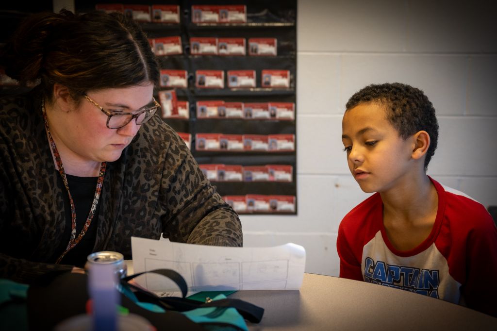 A teacher in a leopard-print cardigan reviews paperwork with a student wearing a red "Captain" jersey in a classroom setting.