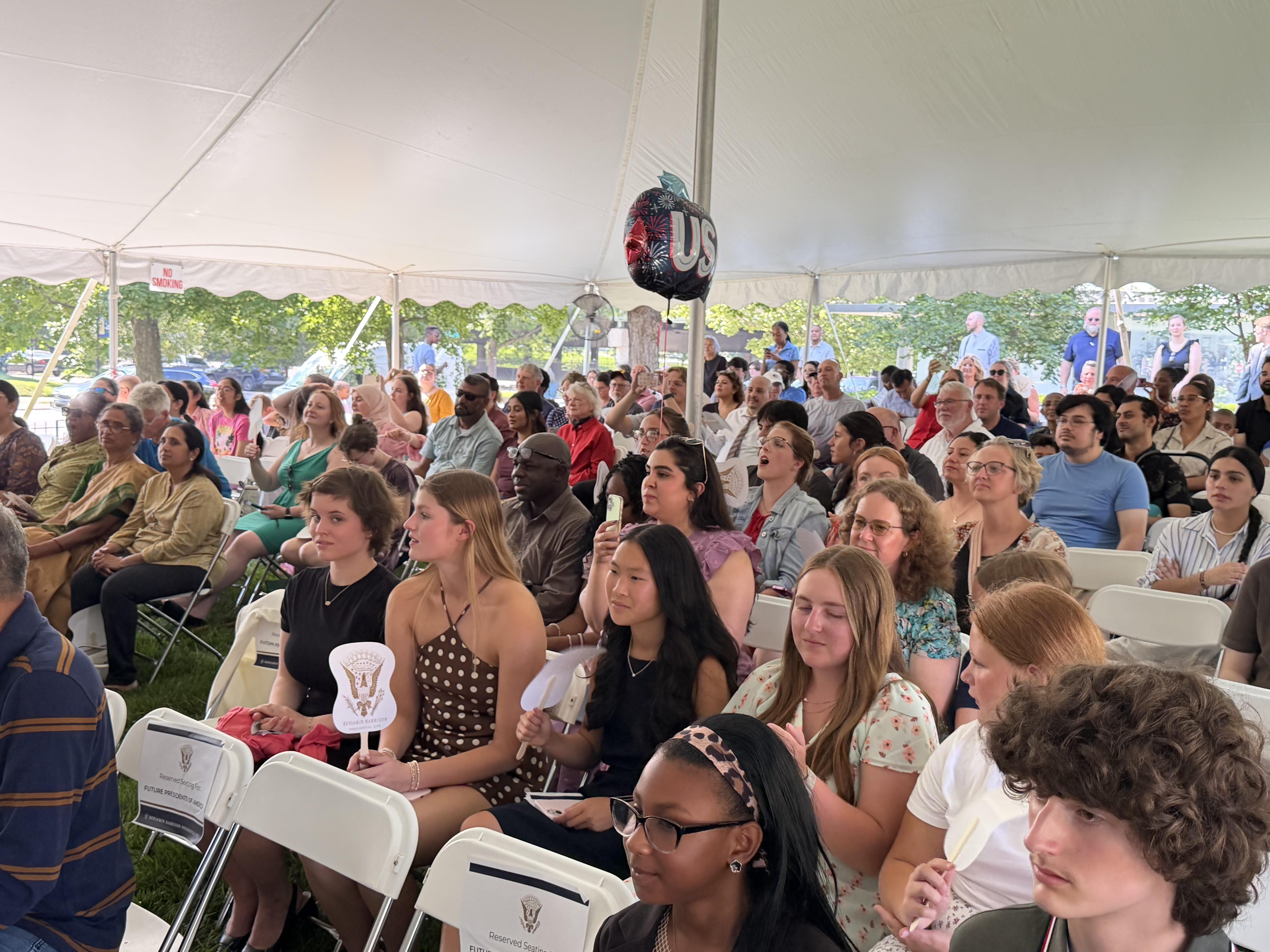 Crowded outdoor event with people seated in white chairs under a tent, with a large "US" logo visible overhead.