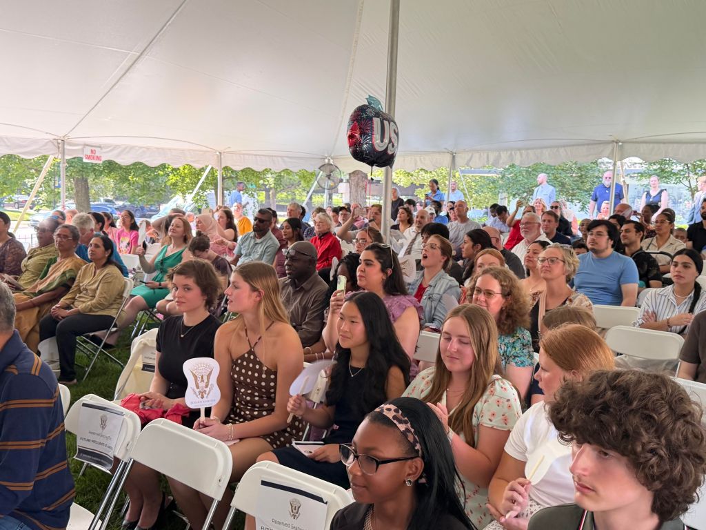 Crowded outdoor event with people seated in white chairs under a tent, with a large "US" logo visible overhead.