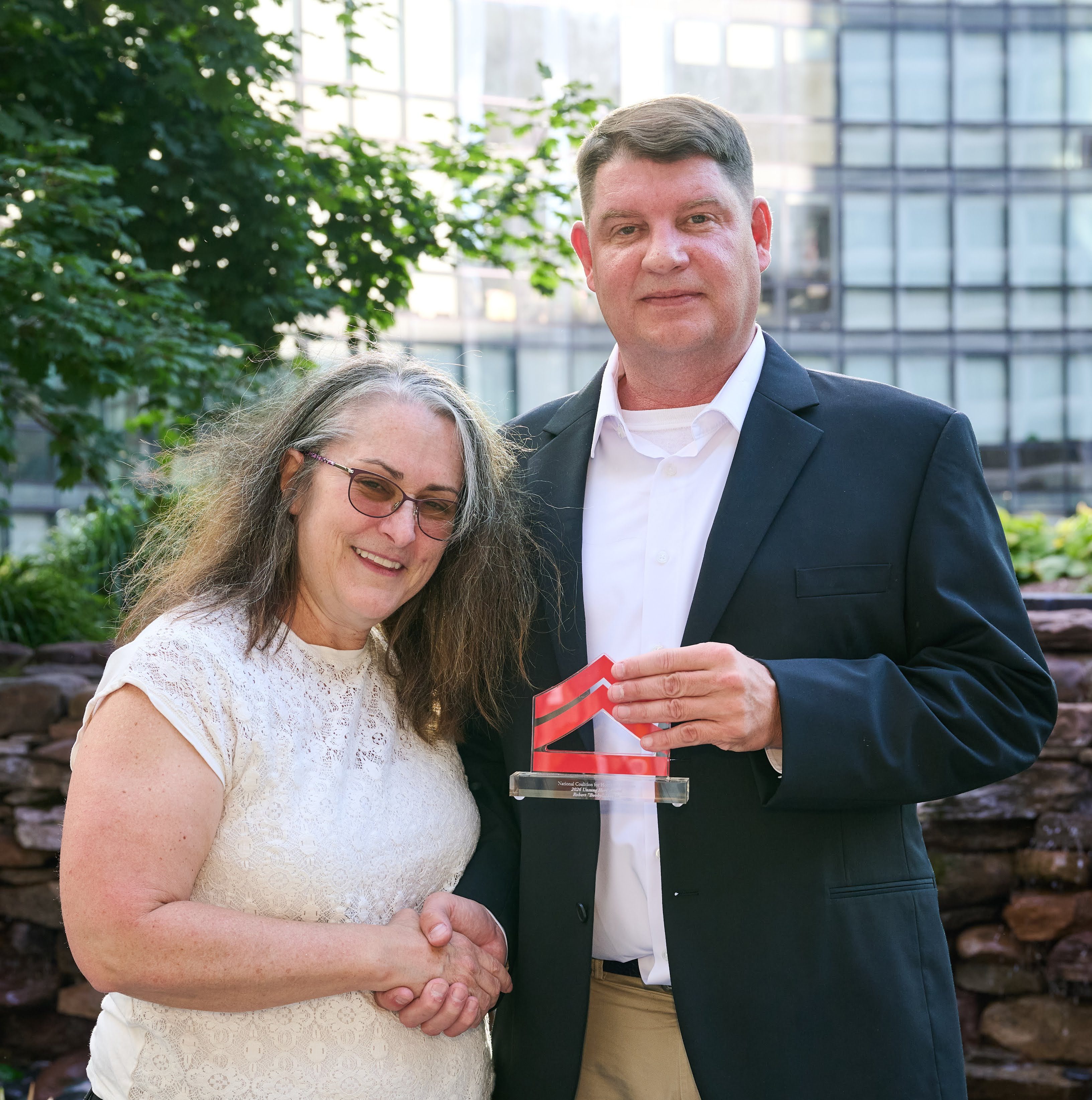 A middle-aged couple standing together outdoors, the woman wearing glasses and the man holding a small award or trophy.