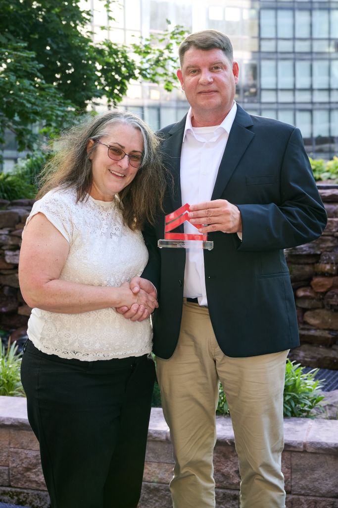 A middle-aged couple standing together outdoors, the woman wearing glasses and the man holding a small award or trophy.