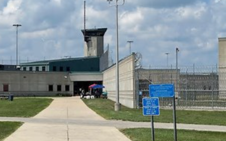Fenced outdoor area with a large building, watchtower, and signage indicating a correctional facility or prison.
