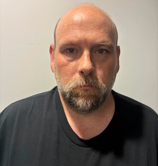 Headshot of a middle-aged man with a bald head and a full beard, wearing a black shirt and looking directly at the camera with a serious expression.