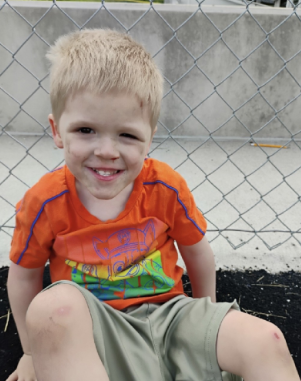 A smiling young boy wearing an orange shirt and green shorts, sitting on a chain-link fence.