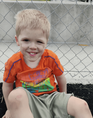 A smiling young boy wearing an orange shirt and green shorts, sitting on a chain-link fence.
