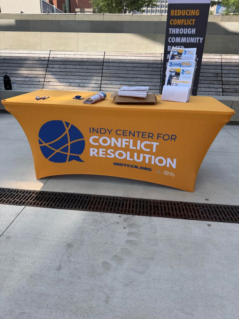 A yellow table with the Indy Center for Conflict Resolution logo and text promoting community-based conflict resolution programs.