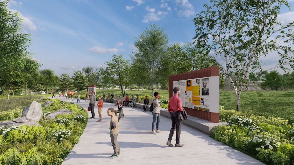 A park scene with people walking on a paved path surrounded by trees and greenery. A display board with portraits and text is visible.