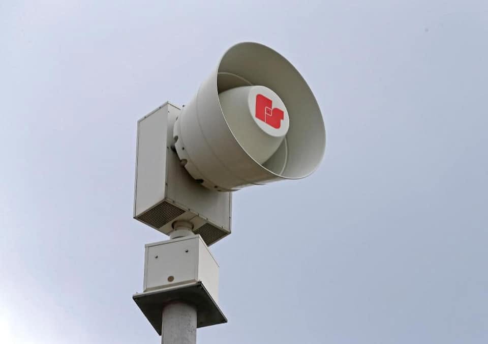 A large gray speaker or siren mounted on a pole, with a red cross symbol visible on the front.