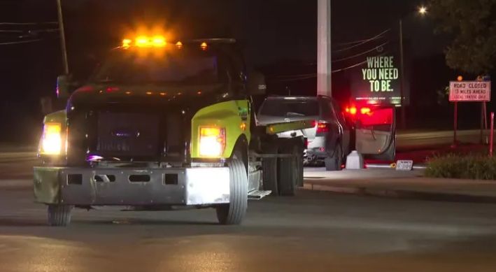Tow truck with flashing lights on a dark street, with a "Where you need to be" sign visible.