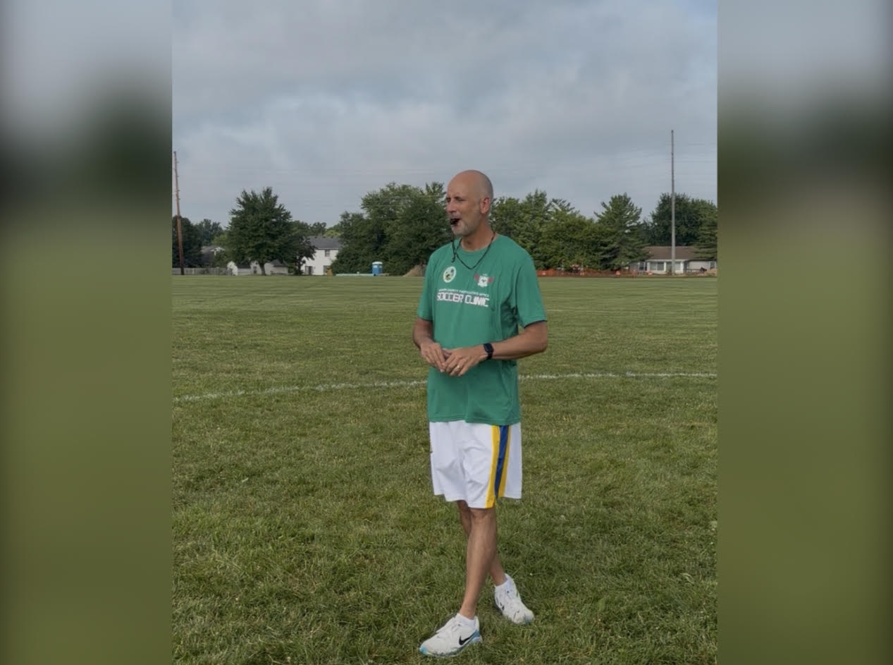 A middle-aged man wearing a green t-shirt with "Soccer Camp" printed on it, standing on a grassy field.