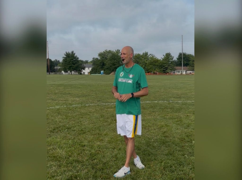A middle-aged man wearing a green t-shirt with "Soccer Camp" printed on it, standing on a grassy field.